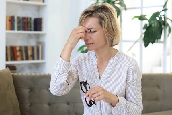 Stressed middle aged woman sit on sofa in living room, lost in thought