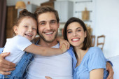 Pozitivní přátelští mladí rodiče s usměvavou dceruškou sedí spolu na pohovce a relaxují doma o víkendu