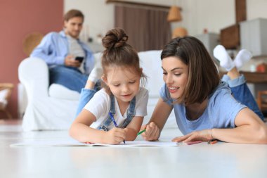 Mutlu anne gülümseyen kız sıcak zeminde uzanıyor yaratıcı aktivitelerin tadını çıkarıyor, albümle resim çiziyor, baba kanepede dinleniyor, aile birlikte boş zaman geçiriyor.