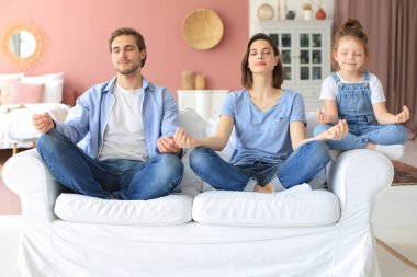 Küçük kızıyla sakin genç bir aile, birlikte kanepede yoga yapar, küçük bir anaokulu çocuğuyla mutlu ebeveynler, kanepeye dinlenir, hafta sonları evde negatif duygulardan arınırlar.