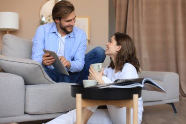 Oturma odasında oturmuş, medya içeriğini internetten izleyerek tablette online alışveriş yapan mutlu genç bir çift.