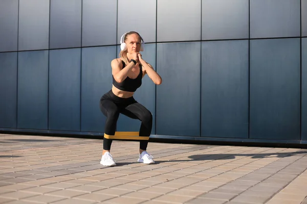 Sporty woman doing squats with fitness gum expander outdoors