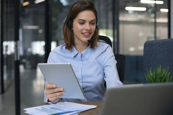 Cheerful female manager sitting at office desk and performing corporate tasks using wireless connection on digital gadgets