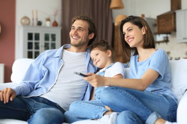 Mutlu ve çocuklu bir aile kanepede oturup televizyon seyrediyor, genç ebeveynler kızını kucaklıyor ve birlikte kanepede dinleniyorlar.