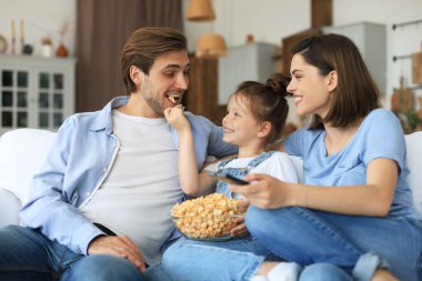 Mutlu ve çocuklu bir aile kanepede oturur televizyon izler ve patlamış mısır yer, genç ebeveynler kızlarını kucaklar birlikte kanepede dinlenirler.