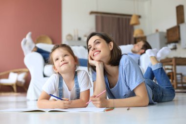 Mutlu anne gülümseyen kız sıcak zeminde uzanıyor yaratıcı aktivitelerin tadını çıkarıyor, albümle resim çiziyor, baba kanepede dinleniyor, aile birlikte boş zaman geçiriyor.