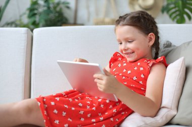 Gülümseyen küçük kız kanepede uzanmış online oyunlar oynuyor, web sörfü bilgileri, tablet üzerindeki komik uygulamaları kullanıyor.