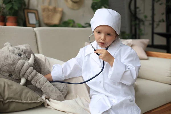 可爱的孩子穿着医生的衣服玩玩具大象 用听诊器检查它的呼吸 — 图库照片