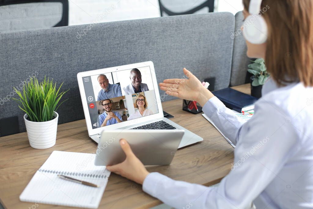 Businesswoman in headphones talking to her colleagues in video conference. Multiethnic business team working from office using laptop, discussing financial report of their company on tablet.