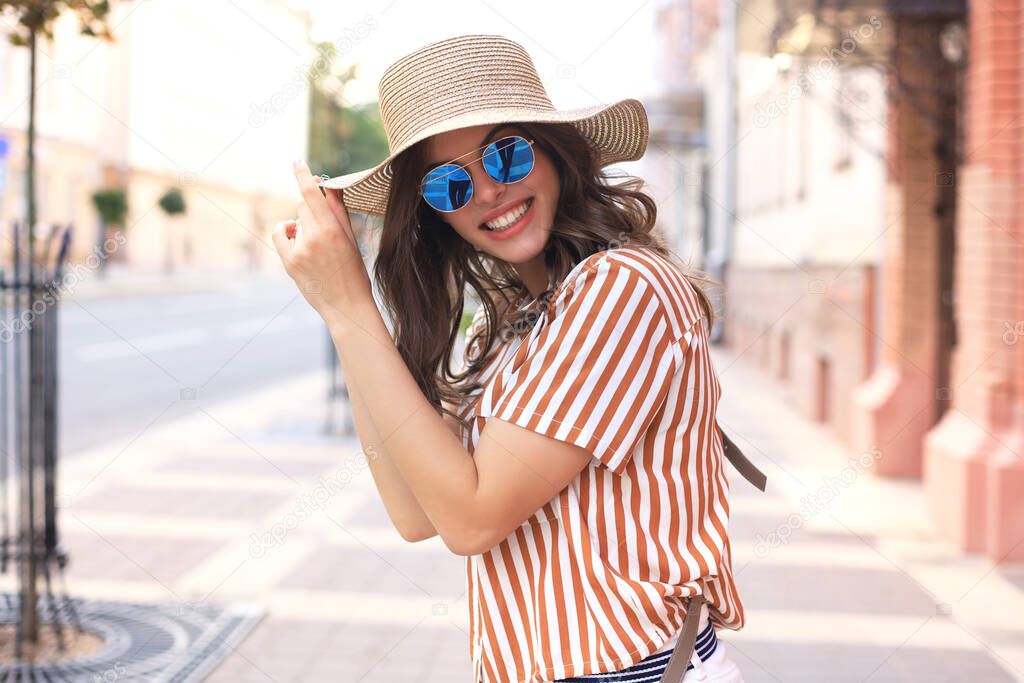 Smiling hipster trendy girl posing at the city summer street,holding hat