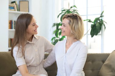 Güzel orta yaşlı anne ve yetişkin kızı evde kanepede otururken sarılıp gülümsüyorlar.