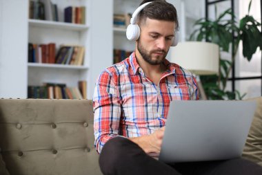 Konsantre olmuş genç serbest çalışan işadamı dizüstü bilgisayarla kanepede oturuyor, uzaktan evde çalışıyor.