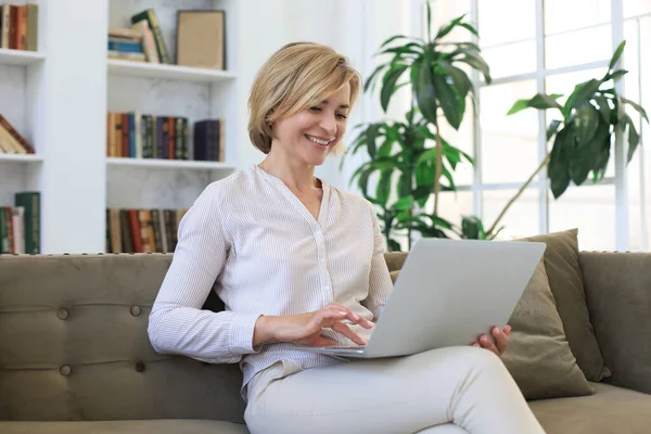快乐的中年妇女坐在沙发上 一边用笔记本电脑 — 图库照片