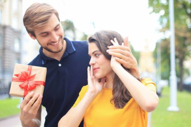 Yazlık giysiler içinde, gülümseyen ve hediye kutusunu açık havada tutan büyüleyici ve heyecanlı bir çift.