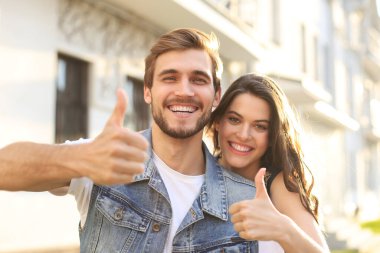 Mutlu, güler yüzlü, genç bir çiftin dışarıda başparmak kaldırarak selfie çektiği bir resim.