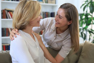 Güzel orta yaşlı anne ve yetişkin kızı evde kanepede otururken sarılıp gülümsüyorlar.
