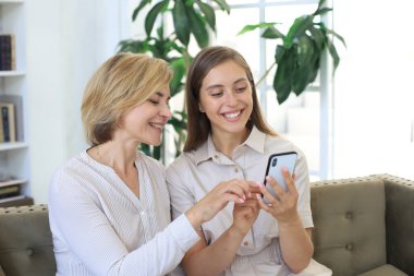 Orta yaşlı anne ve kız kucaklaşıyorlar, birlikte telefon kullanıyorlar, video veya fotoğraf izliyorlar, evdeki rahat kanepede oturuyorlar.