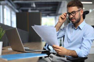 Genç adam ofiste otururken kağıtlarla çalışıyor. Başarılı girişimci dikkatli ve yoğun bakışla belgeleri inceliyor..