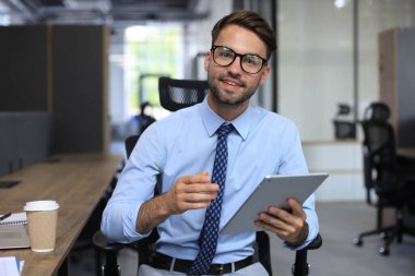 Yakışıklı genç işadamı dijital tablet kullanıyor ve ofiste otururken kameraya bakıyor.