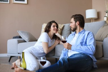 Mutlu çift oturma odasında oturmuş, yerde dinleniyor, kırmızı şarap içiyorlar. Gülümseyen genç karı koca evde dinleniyor. Aile hafta sonlarında romantik buluşmaların tadını çıkarın..