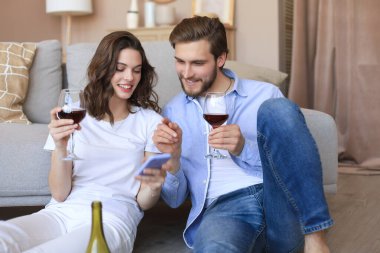 Mutlu çift oturma odasında oturmuş, yerde dinleniyor, kırmızı şarap içiyor, birlikte akıllı telefona bakıyorlar. Gülümseyen genç karı koca evde dinleniyor. Aile hafta sonlarında romantik buluşmaların tadını çıkarın.