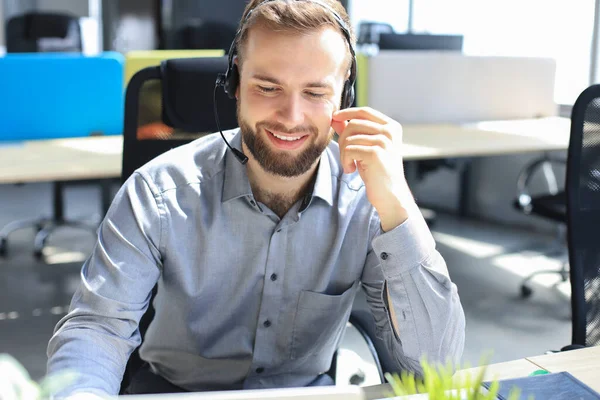 在现代办公室里用耳机对男性呼叫中心操作员微笑 在笔记本电脑上查阅在线信息 在档案中查找信息 以便为客户提供帮助 — 图库照片