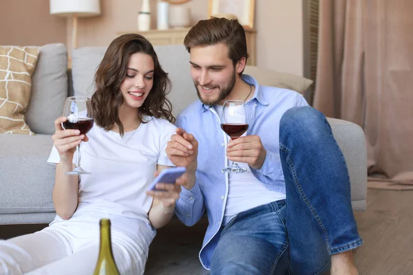 Mutlu çift oturma odasında oturmuş, yerde dinleniyor, kırmızı şarap içiyor, birlikte akıllı telefona bakıyorlar. Gülümseyen genç karı koca evde dinleniyor. Aile hafta sonlarında romantik buluşmaların tadını çıkarın.