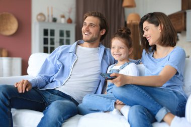 Mutlu ve çocuklu bir aile kanepede oturup televizyon seyrediyor, genç ebeveynler kızını kucaklıyor ve birlikte kanepede dinleniyorlar.