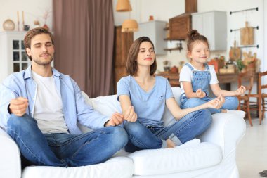 Küçük kızıyla sakin genç bir aile, birlikte kanepede yoga yapar, küçük bir anaokulu çocuğuyla mutlu ebeveynler, kanepeye dinlenir, hafta sonları evde negatif duygulardan arınırlar.