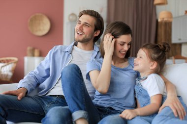 Hafta sonları evde dinlenirken, kanepede birlikte oturan gülümseyen küçük bir kızı olan olumlu, arkadaş canlısı ebeveynler.