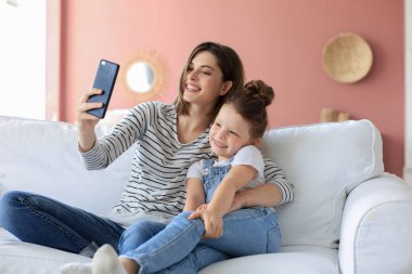 Güzel genç anne ve küçük kızı evde kanepede akıllı telefonlarıyla selfie çekiyorlar.