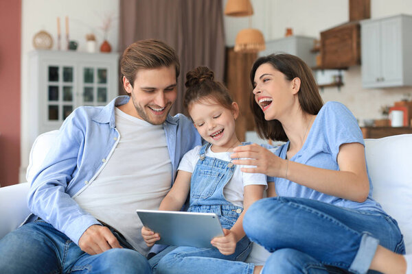Позитивные дружелюбные молодые родители с улыбающейся маленькой дочерью, сидящей на диване вместе, отвечая на видео-звонок на цифровом планшете, расслабляясь дома в выходные