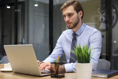 Genç, modern iş adamı ofiste otururken dizüstü bilgisayar kullanıyor.