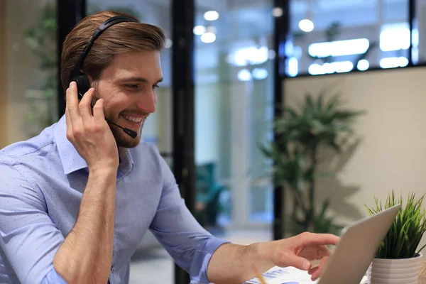 Smiling male business consultant with headphones sitting at modern office, video call looking at laptop screen. Man customer service support agent helpline talking online chat
