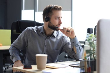 Kulaklıklı, gülümseyen bir erkek çağrı merkezi operatörü, modern ofiste oturuyor, bir dizüstü bilgisayardaki çevrimiçi bilgilere danışıyor, müşteriye yardımcı olmak için bir dosyada bilgilere bakıyor.