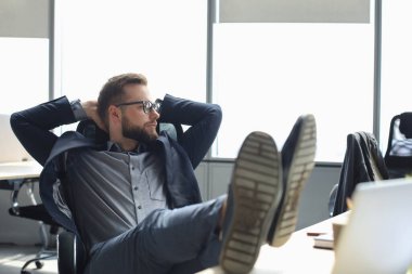 Genç ve modern iş adamı ofiste otururken ellerini başının arkasında tutuyor ve gülümsüyor.