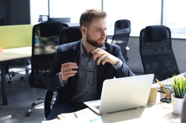 Genç, modern iş adamı ofiste otururken dizüstü bilgisayar kullanıyor.