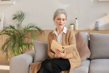 Çekici orta yaşlı kadın oturma odasındaki kanepede oturmuş kitap okurken gülümsüyor.