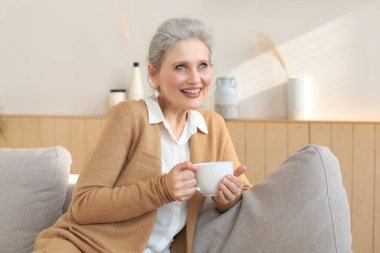 Mutlu olgun bir kadın rahat koltukta dinleniyor kahve ya da çay içiyor, başka tarafa bakıyor, evdeki rahat koltukta dinleniyor sıcak içeceğin tadını çıkarıyor.
