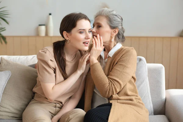Happy middle mother whispering secret to her smiling daughter at home, gossiping, sharing secret. Beautiful woman hearing good news from mature mum, trusted relations
