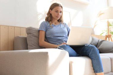 Gülümseyen kız kanepede oturup dizüstü bilgisayarda webinar izliyor. Mutlu genç kadın çevrimiçi uzak kurslarda okuyor.