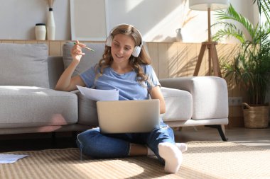 Gülümseyen kız kanepede oturup dizüstü bilgisayarda webinar izliyor. Mutlu genç kadın çevrimiçi uzak kurslarda okuyor.