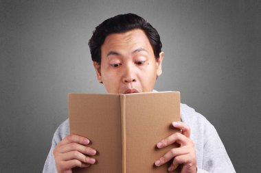 Beyaz gömlek ve ağzını bir kitapla kapsayan gri ceket giyen genç Asyalı adam ifade şaşırttı. Arka plan gri. Baş ve omuzlar kapatın