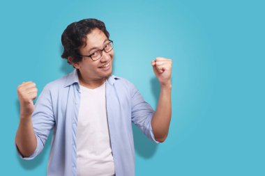 Küçük Asya adam giyen beyaz ve mavi gömlek mutlu ifade, zafer jest kutluyor. Beyaz izole. Vücut portre kadar kapatın