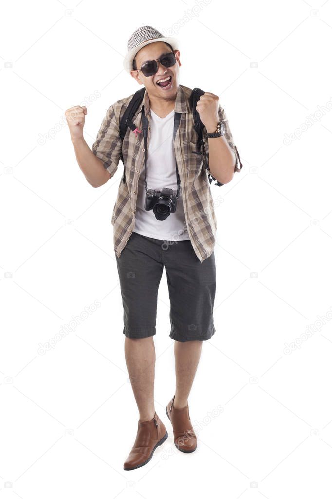 Traveling people concept. Portrait of Asian male backpacker tourist wearing hat, black sunglasses, camera and backpack isolated on white. Full body portrait showing surprised happy winning gesture