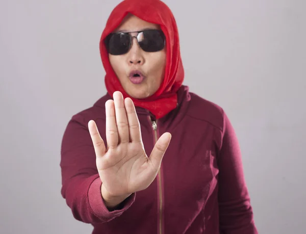 Portrait of muslim lady wearing black sunglasses in red suit and hijab shows stop sign, prohibit forbid disallow something bad with palm hand gesture