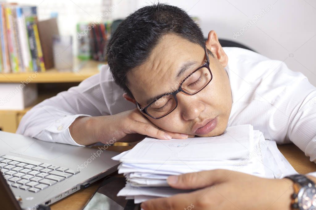 Portrait of Asian businessman tired for overworked and sleep in front of his laptop and paperworks at the office