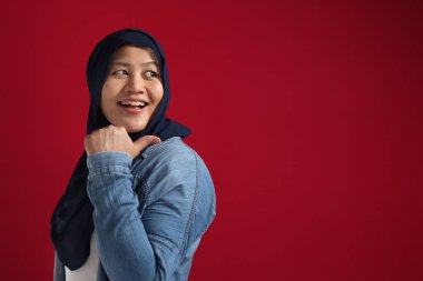 Asyalı genç, mutlu Müslüman kadının portresi kırmızı arka planda fotokopi alanı olan bir şeyi kendi tarafında sunduğunu gösteriyor.