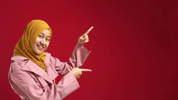 魅力的な美しいイスラム教徒の十代の少女の肖像画を身に着けているヒジャーブ笑顔とコピースペースで側面を指す 何かを提示 赤の背景に対するマーケティングの概念 — ストック写真