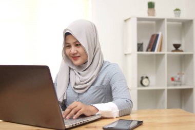 Evdeki bilgisayarda çalışan, tesettürlü, Asyalı, genç, özgür bir müslüman kadın. Çevrimiçi öğrenim gören çekici iş kadını, dizüstü bilgisayar, web sörfü bilgileri veya internet mağazasından alışveriş yapıyor.